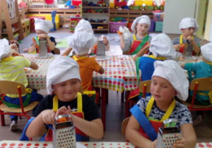 Grupa dzieci przy swych stolikach w strojach kucharskich ścierająca marchewki na tarkach.